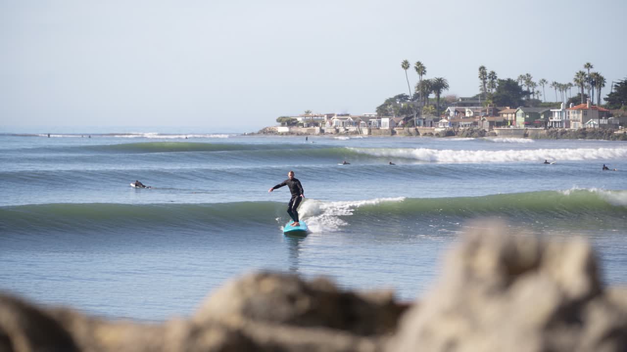 AI illustration of an offshore surfer on a blue soft-top board, viewed past foreground rocks